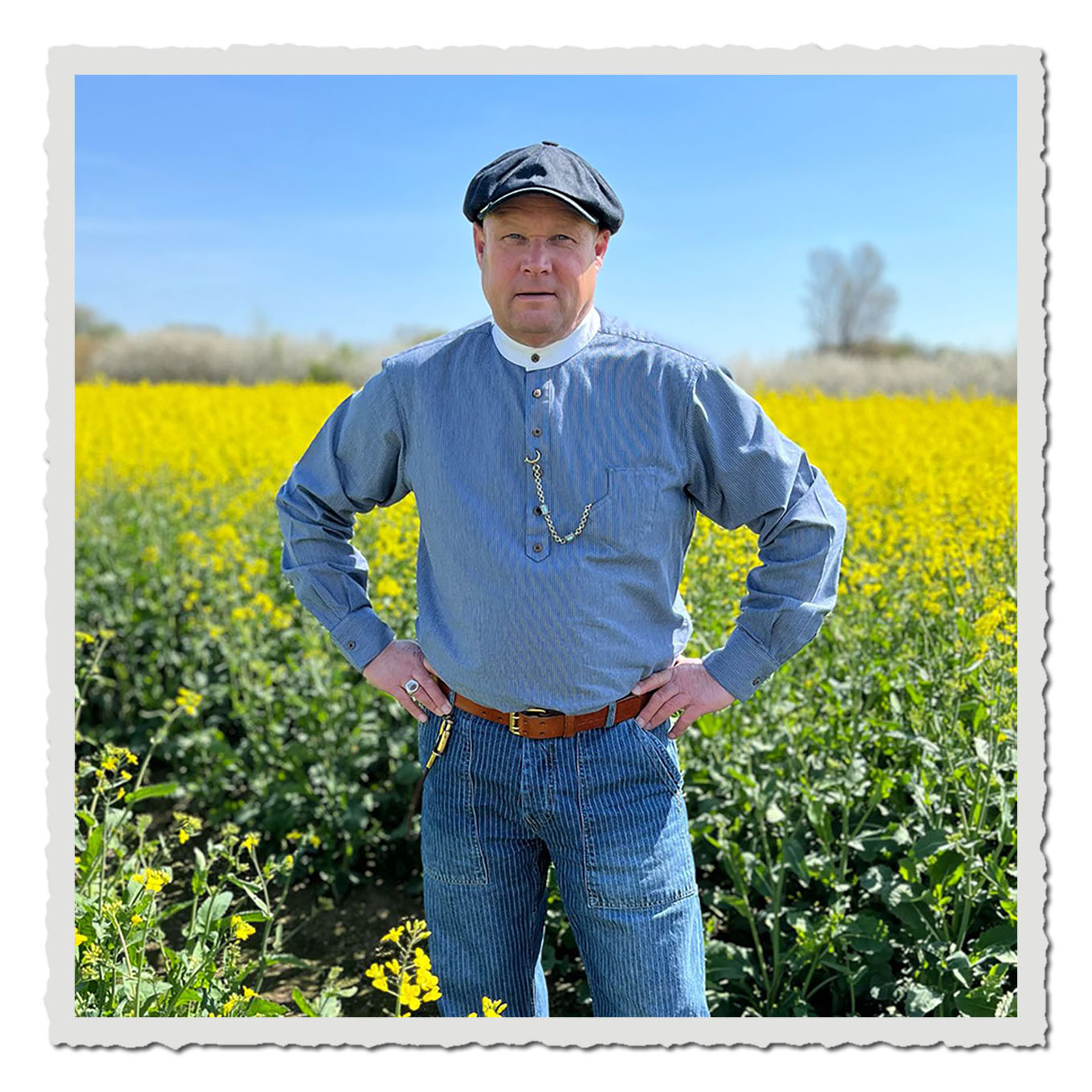 Person im blau gestreiften Stehkragenhemd auf einem gelben Rapsfeld unter blauem Himmel.
